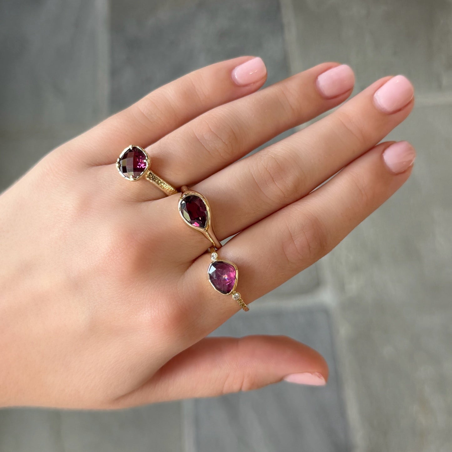 A close-up of a hand wearing three gold rings, each featuring a teardrop-shaped deep red gemstone, set against a blurred gray tiled background.