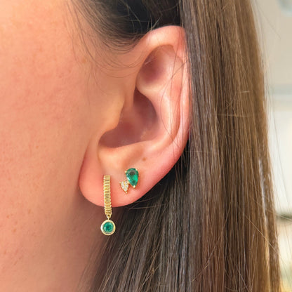 Close-up of a person's ear wearing a gold huggie hoop earring with a dangling emerald and a small emerald and diamond stud in the lobe.
