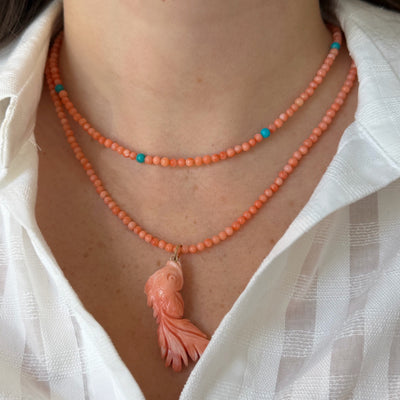 A close-up of a person's neck and upper chest wearing a double-strand coral necklace with turquoise accent beads and a carved coral fish pendant, paired with a white button-down shirt.