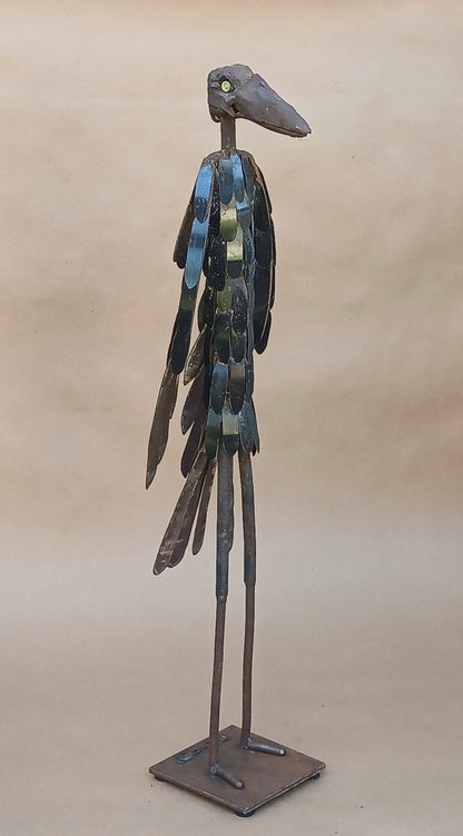 A tall metal bird sculpture with a long beak and long legs, standing on a square base against a plain beige background.